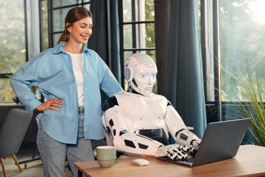 Young woman with a cup of coffee and a humanoid robot working while sitting at a laptop in a modern office. Collaboration between humans and artificial intelligence.